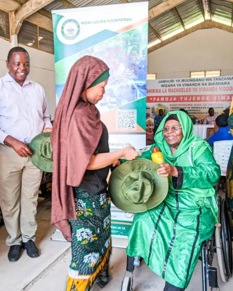 A woman in a green dress, seated in a wheelchair, receives a hat from another woman at a Riziki Lulida Foundation event. A foundation banner stands behind them.
