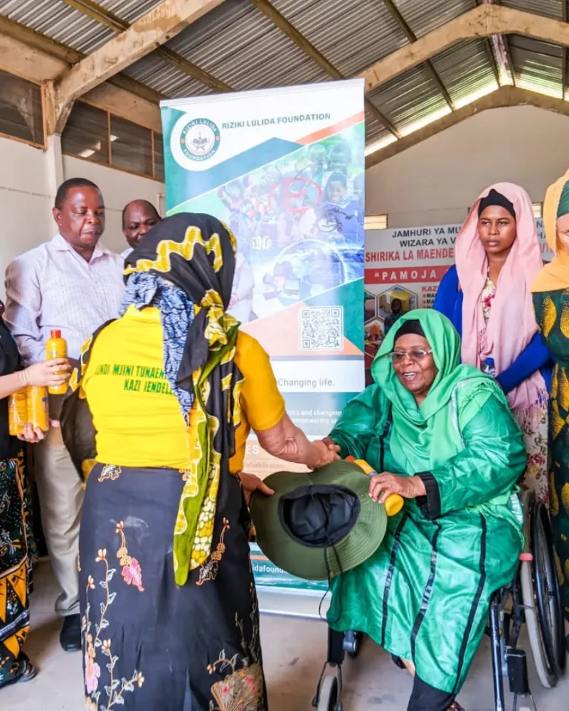 A group of women and men gather around a woman in a wheelchair at a Riziki Lulida Foundation event. Some of them hold bottles.