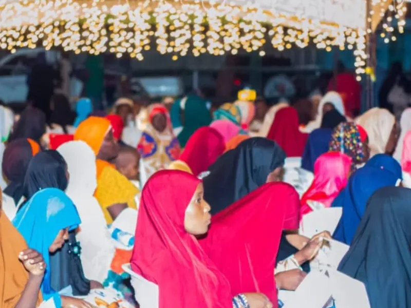 Women and girls in colourful headscarves sit in an audience under strings of lights at an evening event.