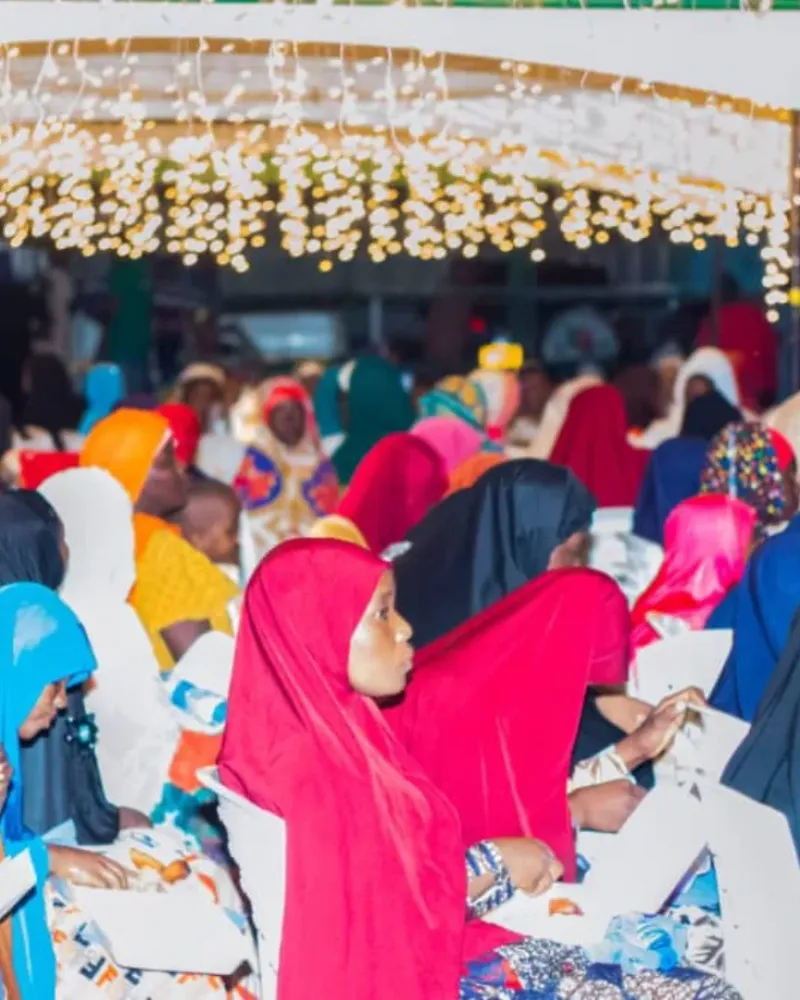 Women and girls in colourful headscarves sit in an audience under strings of lights at an evening event.