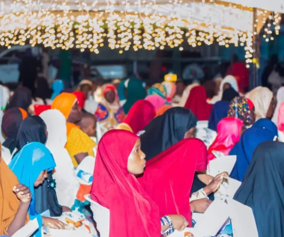 Women and girls in colourful headscarves sit in an audience under strings of lights at an evening event.