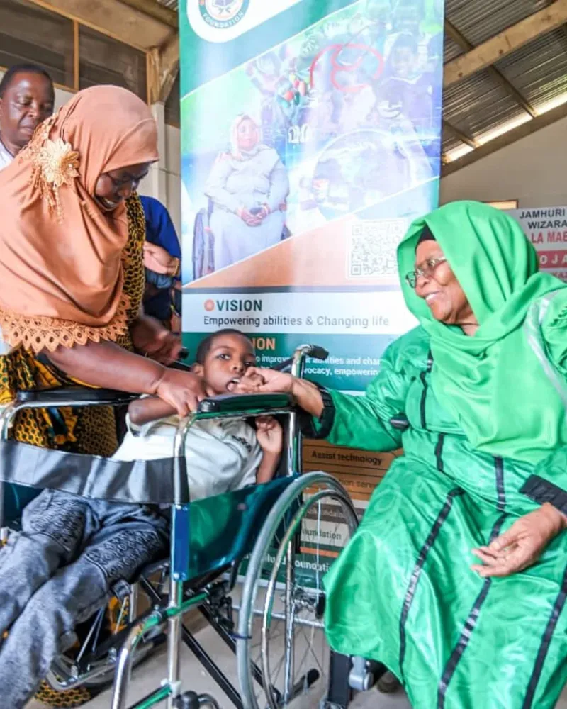 A woman in a green dress, seated in a wheelchair, reaches out to a child in a wheelchair. A woman beside the child holds the chair.